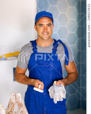 Professional man builder in blue construction overalls and cap posing with gloves and trowel in apartment Professional man builder in blue construction overalls and cap posing with gloves and trowel in apartment 106625632