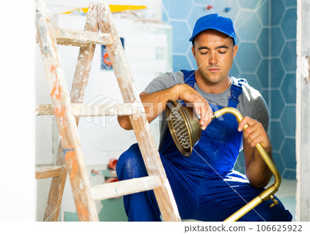 Skilled workman wearing in blue construction overalls sitting squat and repairing shower head in bathroom Skilled workman wearing in blue construction overalls sitting squat and repairing shower head in bathroom 106625922
