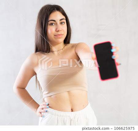 Young woman showing her mobile phone standing against wall 106625975