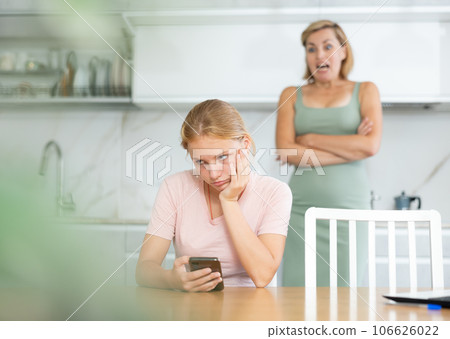 Upset teenage girl sitting with phone while disapproving mother standing in background 106626022