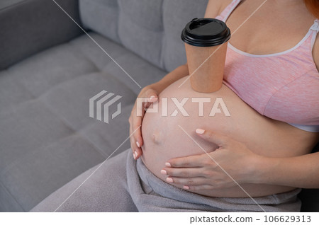 Faceless pregnant woman in 3rd trimester holding a coffee glass on her belly.  106629313