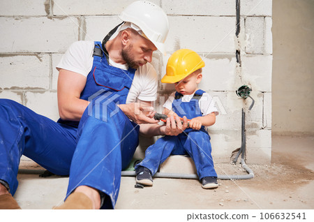 Male builder showing wire stripper cutter tool to child. Man and kid in safety helmets and work overalls sitting by brick wall in building under construction. Home renovation and parenting concept. Male builder showing wire stripper cutter tool to child. Man and kid in safety helmets and work overalls sitting by brick wall in building under construction. Home renovation and parenting concept. 106632541