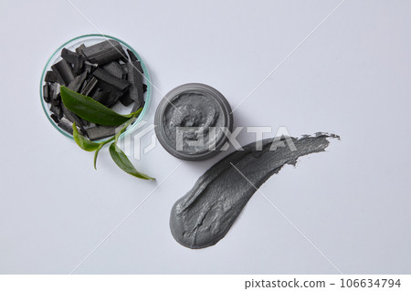 On the petri dish are bamboo charcoal and green tea bud decorated with a black jar container mask and streaks of mask substance on a light gray background. Top view, advertising photo 106634794