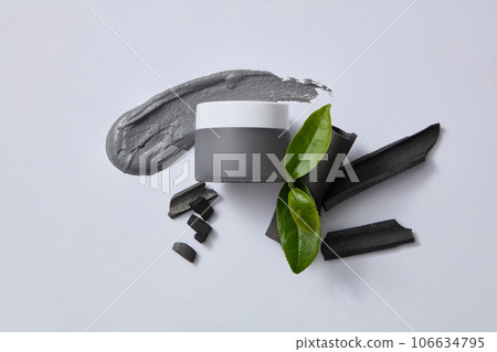 A jar container cream or mask decorated with bamboo charcoal and tea leaves on light gray background. Extracted from green tea and bamboo charcoal help the skin fight bacteria and remove oil  106634795
