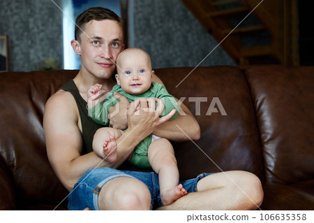 Young man father parent sits on leather sofa with his six month old baby. 106635358