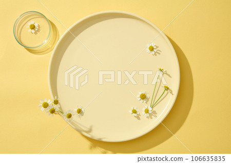 On the yellow background, a round plate decorated with chamomilla (Tanacetum parthenium). Minimal concept with copy space for cosmetic of chamomilla extract presentation On the yellow background, a round plate decorated with chamomilla (Tanacetum parthenium). Minimal concept with copy space for cosmetic of chamomilla extract presentation 106635835