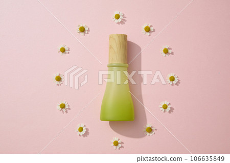 A green glass bottles with wooden cap displayed on a round shape of chamomilla flowers on a pink background. Scene for advertising and cosmetics product presentation A green glass bottles with wooden cap displayed on a round shape of chamomilla flowers on a pink background. Scene for advertising and cosmetics product presentation 106635849