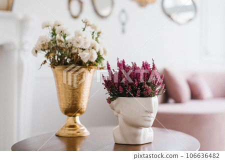 Purple heather at the vase sculpture of a head and white chrysanthemums in a golden classical vase on a table in the interior of a cozy house 106636482
