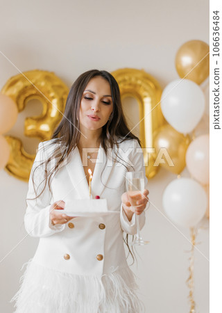 Positive woman celebrating anniversary dressed in stylish feather dress blowing out candle on birthday cake around inflated balloons at home 106636484
