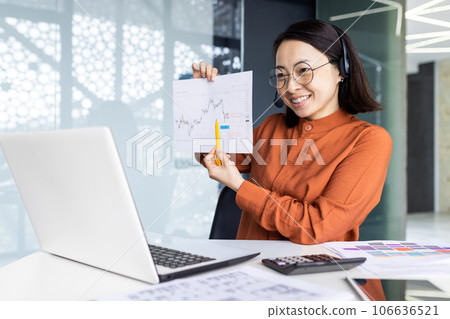 Online presentation remote meeting, asian woman with headset phone smiling and showing to laptop camera graph for good and successful achievement results, female worker inside office. Online presentation remote meeting, asian woman with headset phone smiling and showing to laptop camera graph for good and successful achievement results, female worker inside office. 106636521