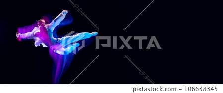 Young guy in stylish white clothes in motion, jumping against black studio background in neon with mixed lights effect. Banner 106638345