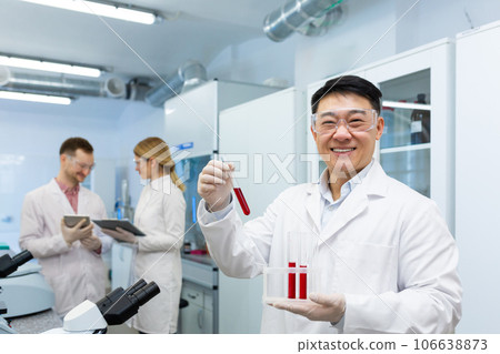 Portrait of a smiling young doctor of Asian descent standing in the laboratory and holding flasks with blood samples. Colleagues are discussing work behind. 106638873