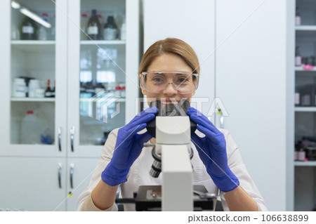 Close-up photo of a young woman working in a laboratory under a microscope, sitting at a table in a uniform and glasses and smiling at the camera. 106638899