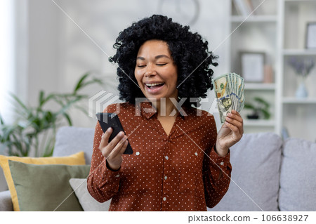 African american young smiling woman sitting on sofa at home using mobile phone and slurping money cash in hand. African american young smiling woman sitting on sofa at home using mobile phone and slurping money cash in hand. 106638927