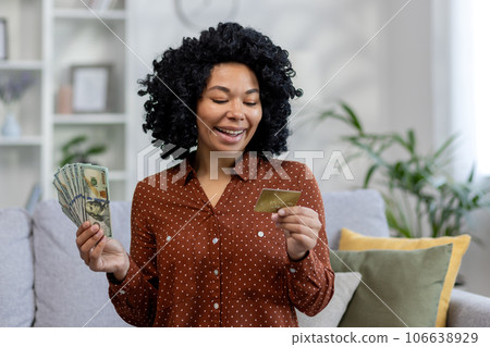 Smiling african american woman holding cash banknotes and looking at credit card. Sitting at home on the couch. 106638929