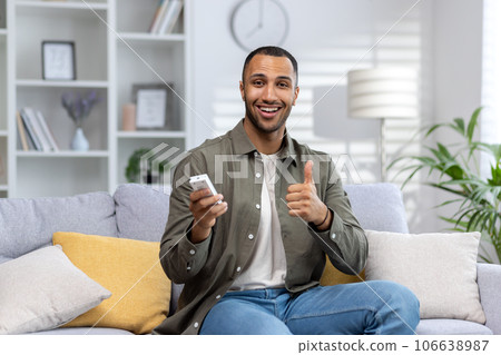 Portrait of a young African American man sitting on the sofa at home and holding the air conditioner remote control, smiling at the camera and showing the super gesture with his finger. 106638987