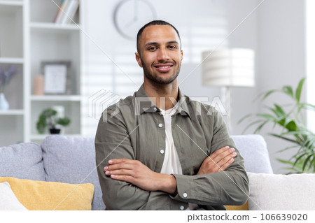 Portrait of a young Muslim man sitting on the couch at home and looking at the camera smiling, arms crossed. 106639020