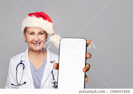 Christmas portrait of smiling woman doctor in Santa hat showing smartphone with blank emptydisplay Christmas portrait of smiling woman doctor in Santa hat showing smartphone with blank emptydisplay 106645408