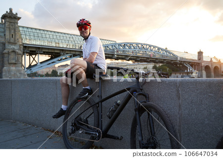 A male cyclist sits next to a bicycle on the embankment, resting after a bike ride A male cyclist sits next to a bicycle on the embankment, resting after a bike ride 106647034