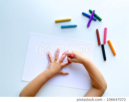 Childrens hands draw their hand with wax crayons on white paper, top view. Childrens hands draw their hand with wax crayons on white paper, top view. 106647072