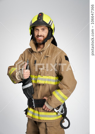 Professional firefighter showing thumb up to camera in studio. Portrait of handsome bearded fireman in uniform gesturing, giving like and smiling, on gray background. Concept of work, hand gesture. 106647366