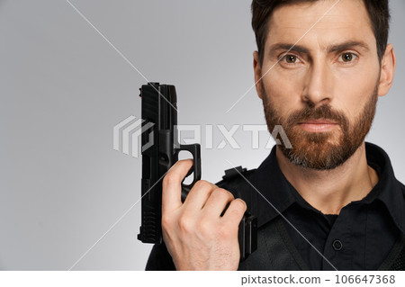Serious officer keeping short-barrelled, firearm gun close to face, while looking at camera. Close up portrait of policeman, armed with handgun, on gray background. Concept of profession, weapon. 106647368