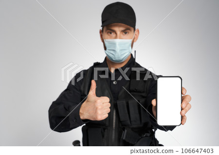 Male police officer in protective mask demonstrating phone's screen, with empty space. Portrait of policeman showing thumb up, looking camera, isolated on gray. Concept of safety, call, quarantine. 106647403