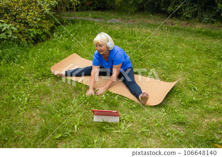 Athletic 50s senior woman uses digital tablet during workout watches online session or workout program poses at yoga mat outdoors dressed in sport clothes alone in the park or forest in nature Athletic 50s senior woman uses digital tablet during workout watches online session or workout program poses at yoga mat outdoors dressed in sport clothes alone in the park or forest in nature 106648140