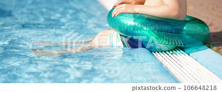 Happy little boy child siting on the edge of the pool with green inflatable ring. Kids legs dipping in the water of the blue swimming pool on sunny day at summer vacation. Banner with copy space Happy little boy child siting on the edge of the pool with green inflatable ring. Kids legs dipping in the water of the blue swimming pool on sunny day at summer vacation. Banner with copy space 106648218