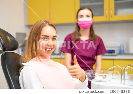 Young woman patient showing thumb up sitting in medical chair at dental clinic. Stomatologist standing behind the patient Young woman patient showing thumb up sitting in medical chair at dental clinic. Stomatologist standing behind the patient 106648288