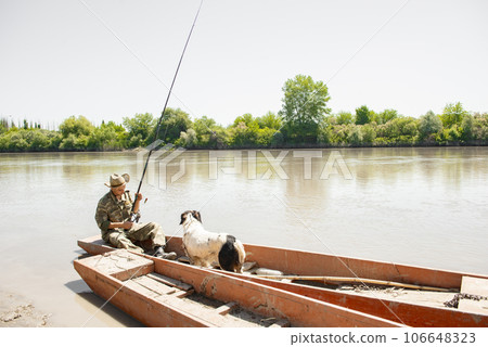 Focused male angler fiddling with tackle, while sitting in shabby boat on shore. Panoramic view of fisherman with cute dog, fishing, with calm spreading river on background. Concept of pastime. 106648323