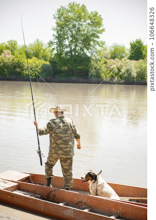Slim male angler in uniform outfit standing in shabby boat with rod, angling in morning. Back view of unrecognizable fisherman going fishing, while tired dog sitting next. Concept of resting together. Slim male angler in uniform outfit standing in shabby boat with rod, angling in morning. Back view of unrecognizable fisherman going fishing, while tired dog sitting next. Concept of resting together. 106648326