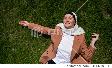 Top view of young woman dressed in hijab and jacket lying on green lawn.  106648391