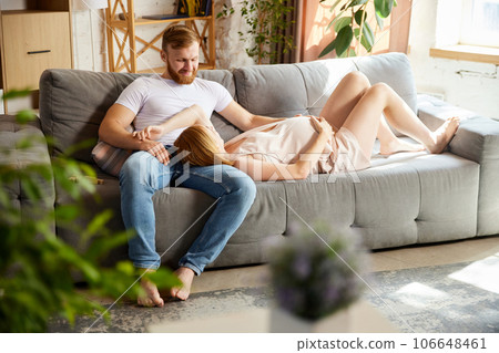 Coziness and care. Husband spending time with his beautiful pregnant wife at home. Woman lying on man's leg Coziness and care. Husband spending time with his beautiful pregnant wife at home. Woman lying on man's leg 106648461