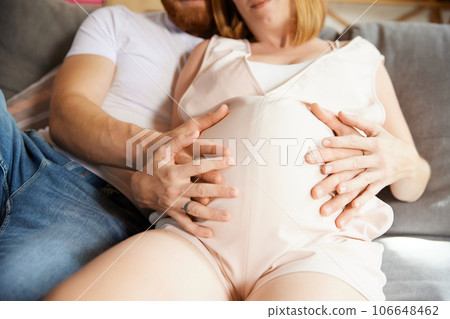 Cropped close-up image of couple sitting and hugging. Man holding hand on his pregnant wife belly Cropped close-up image of couple sitting and hugging. Man holding hand on his pregnant wife belly 106648462