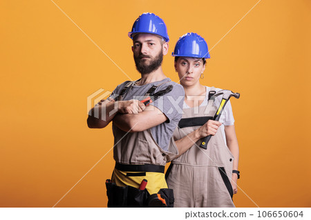 Confident professional renovators posing with pair of pliers and hammer, holding slegdehammer and renovating tools. Man and woman working as constructors with overalls and helmet. 106650604
