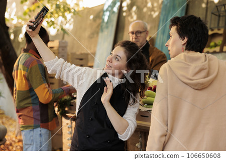Happy woman farmer holding mobile phone making photo with customer while selling organic produce at local street market. Female vegetable stall owner promoting her farming business on social media 106650608