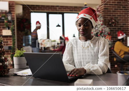 African american woman company manager wearing santa claus hat typing on laptop in christmas decorated office. Employee sitting at desk and working on computer in festive workspace 106650617