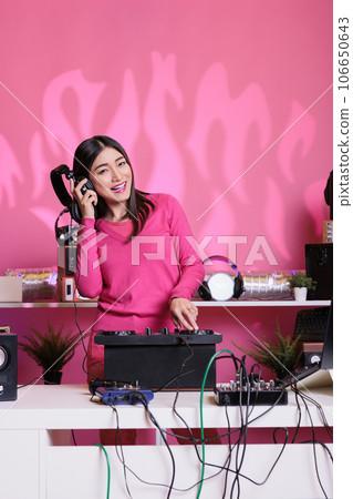 Cheerful performer with headphones playing electronic song at mixer console, standing at dj table having fun with fans at night in club. Artist performing techno music in studio over pink background Cheerful performer with headphones playing electronic song at mixer console, standing at dj table having fun with fans at night in club. Artist performing techno music in studio over pink background 106650643