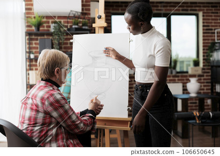 Senior woman engaging in social activities, taking drawing lesson. African American young woman art instructor teaching older adults to draw in community center standing near easel assisting student Senior woman engaging in social activities, taking drawing lesson. African American young woman art instructor teaching older adults to draw in community center standing near easel assisting student 106650645