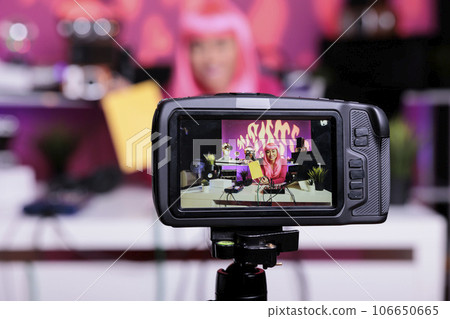 Asian vlogger holding notebook presenting product in front of camera while recording school supplies review using broadcast equipment. Content creator with pink hair live streaming 106650665