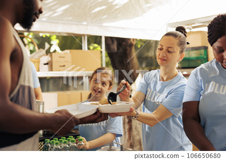 Aid organization recruits a diverse team to distribute free food, supporting less privileged and homeless people, in an effort to alleviate poverty. Volunteer women feed hungry african american man. 106650680