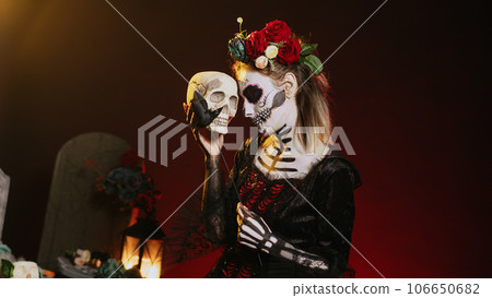 Glamorous woman acting creepy with skull in hand, holding black roses and wearing scary make up. Looking like santa muerte with body art on holy mexican celebration, being flirty. Handheld shot. Glamorous woman acting creepy with skull in hand, holding black roses and wearing scary make up. Looking like santa muerte with body art on holy mexican celebration, being flirty. Handheld shot. 106650682