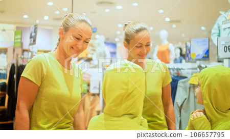 Young mother and child chooses clothes in sports clothing boutique. Cheerful child and mother choosing sports clothes in retail store. Mother and daughter choose looking for clothes in shopping mall. Young mother and child chooses clothes in sports clothing boutique. Cheerful child and mother choosing sports clothes in retail store. Mother and daughter choose looking for clothes in shopping mall. 106657795