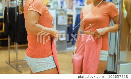 Trendy girl looks in mirror and try outfits in modern shopping centre department store. Happy shopper girl posing in mirror in shopping mall. Sporty girl customer is looking and buying sport clothes. 106657828