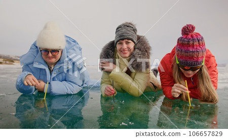 Siberia tradition cocktail Kiss of Baikal is entertainment for tourist on Lake Baikal.Three woman drink alcohol liquid without straw into makeshift glasses in hole frozen ice.Fun vacation trip travel. 106657829
