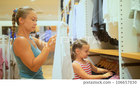 Cute shopaholic mom and daughter choose looking for clothes in clothing store. Mom and daughter shopper customer is looking and buying casual clothes in clothing store. Concept shopping clothes store. Cute shopaholic mom and daughter choose looking for clothes in clothing store. Mom and daughter shopper customer is looking and buying casual clothes in clothing store. Concept shopping clothes store. 106657830
