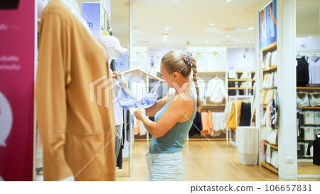 Happy woman looking at clothing in shopping center clothing store mall. Female shopper enjoying purchase in branded fashion boutique in shopping center. Shopper looking at clothing indoors in store. 106657831