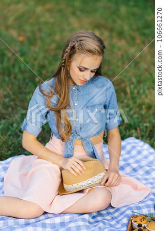 Young woman is sitting on a picnic blanket and holding a straw hat in her hands Young woman is sitting on a picnic blanket and holding a straw hat in her hands 106661270