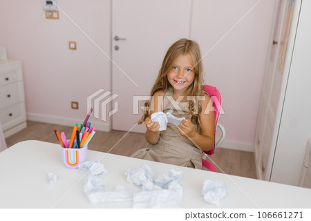 Caucasian schoolgirl is sitting on a chair at a table with a torn page from a school notebook with an incorrectly completed task and laughing Caucasian schoolgirl is sitting on a chair at a table with a torn page from a school notebook with an incorrectly completed task and laughing 106661271